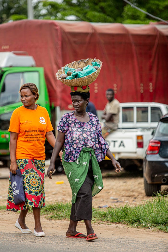 Two African women walking outdoors, one carrying a basket on her head.